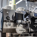 A close-up of a stainless steel espresso machine with a white Sanremo London ceramic cup placed under the portafilter. Stacked paper cups and a glass bean hopper are visible in the background.