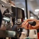 A close-up of a person's hand holding a portafilter under the steam wand of a black and silver espresso machine in a cafe setting.