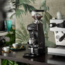 Coffee grinder on a counter with a coffee machine and cups in the background.