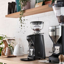 Coffee grinder on a kitchen counter with a white tiled wall and wooden shelf in the background.