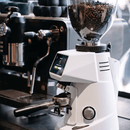 A white commercial coffee grinder filled with dark roasted coffee beans, shown on a dark countertop in a cafe setting with an espresso machine blurred in the background.