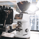 A white electric coffee grinder with a digital display screen and a clear bean hopper filled with coffee beans, set on a counter next to an espresso machine in a cafe environment.