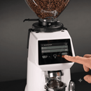 A white electric coffee grinder filled with roasted coffee beans, shown with a person's finger pressing the touchscreen control panel. The grinder has a clear bean hopper at the top and a metal portafilter holder at the bottom, set against a dark gray background.