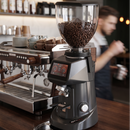 A dark gray electric coffee grinder filled with roasted coffee beans sits on a wooden counter in a cafe setting, with an espresso machine and a blurred barista in the background.