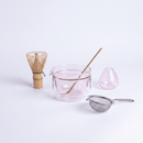 A set of tea-making glassware and accessories on a clean white background, including a pink double-walled bowl with a bamboo whisk inside, a small pink teapot, a metal strainer, and a bamboo whisk.